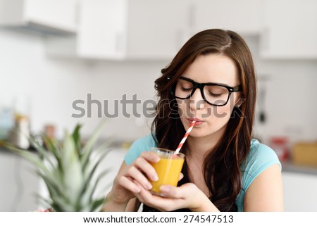 Young woman wearing heavy rimmed glasses sitting enjoying a glass of fresh healthy orange juice in her kitchen