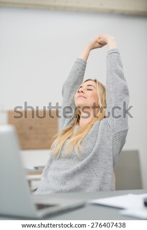 Young businesswoman stretching her arms above her head to relax with a smile of pleasure as she sits at her desk