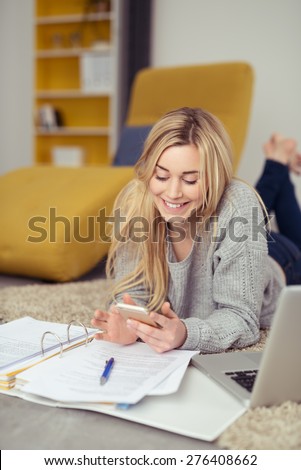 Happy Blond Girl Lying on her Stomach, Using her Mobile Phone While Studying her Lessons with Papers and Laptop on the Floor.