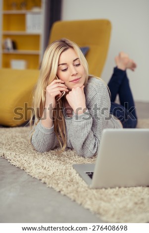 Pretty Blond Girl Lying on her Stomach While Calling Someone on Mobile Phone and Looking at the Laptop on the Floor.