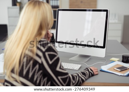 Young blond woman working on a desktop computer at her desk in the office, viewed from behind with a blank white screen