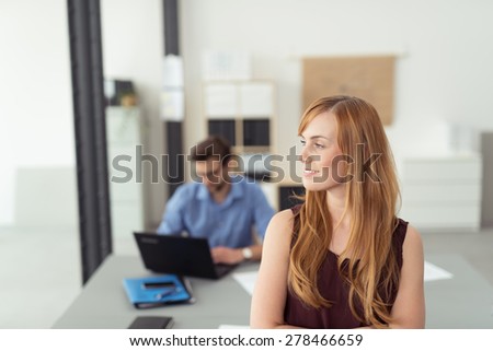 Thoughtful Young Blond Office Woman Leaning Against The Table, Smiling to the Left of the Frame with Busy Male Co-Worker Behind.
