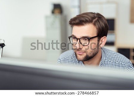 Smiling Young Businessman with Eyeglasses Sitting at his Desk and Facing at the Computer Screen, Captured in Close up.