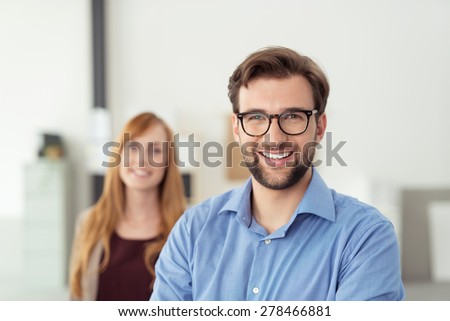 Happy Young Businessman Inside the Office, Wearing Blue Polo Shirt with Eyeglasses, Smiling at the Camera In Front his Female Co-worker.