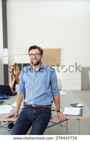 Smiling young man relaxing on the edge of his desk in a large open-plan modern office giving the camera a friendly smile