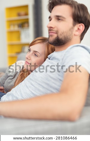 Young husband and wife relaxing together arm in arm on a sofa in the living room watching something to the left off frame, probably television
