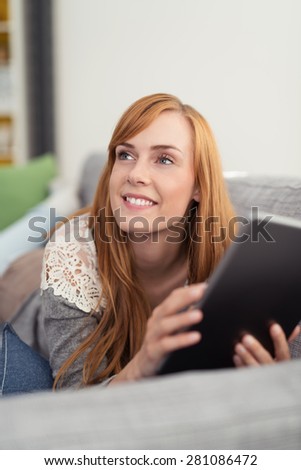 Pretty redhead woman reading a book as she lies on a comfortable sofa looking up with a smile off to the left of the frame