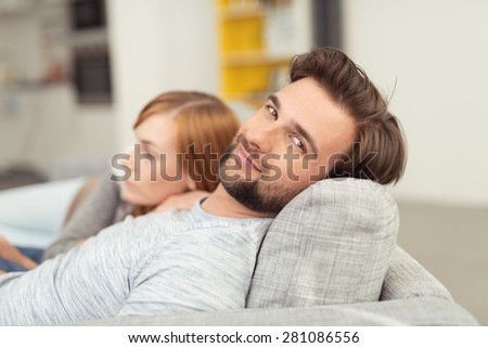 Young Man with Facial Hair Smiling at Camera with Head Leaned Back on Sofa Cushion, Relaxing with Woman Curled Up Beside Him