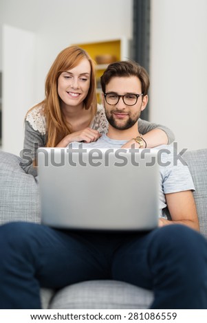 Man with Facial Hair and Glasses Sitting on Sofa Using Laptop Computer with Red Haired Woman Looking Over Shoulder at Screen Leaning Over Back of Sofa