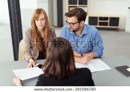 Married couple discussing investments with a broker as they sit together at a desk in her office going through paperwork together, view from behind the agent