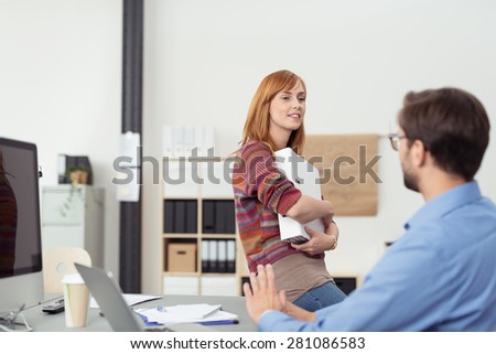 Young woman having an informal meeting with a colleague in the office as she perches on the edge of his desk clutching an office binder