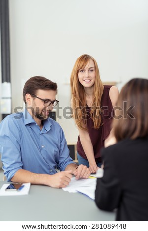 Young couple in a meeting with a broker or agent sitting at her desk looking at paperwork with focus to the smiling wife