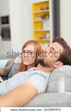 Attractive woman relaxing at home with her husband who is dozing with his head on the back of the sofa as she smiles at the camera, view across his body