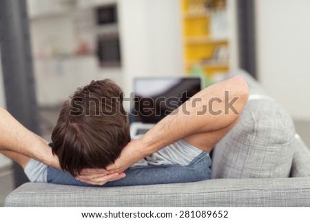 Man Relaxing with Hands Behind Head on Sofa and Laptop Computer on Lap Visible Out of Focus in Distance