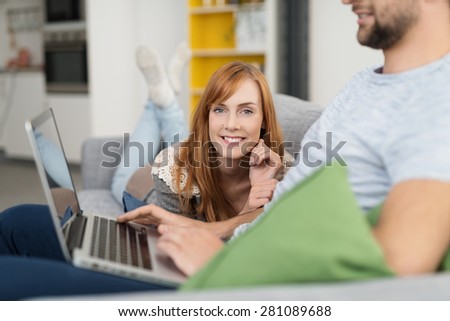 Red Haired Woman Lying on Sofa and Smiling at Camera with Man Typing on Laptop Computer in Foreground
