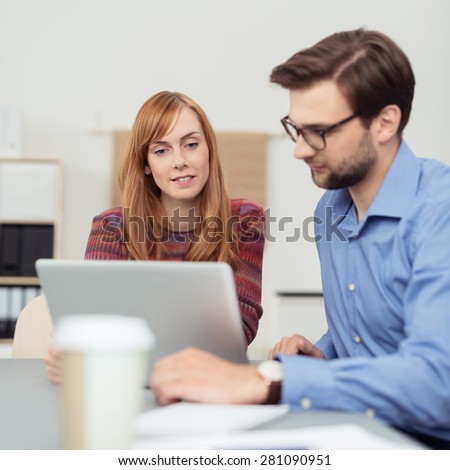 Two colleagues working together at the office with an attractive young woman and man sitting reading a laptop screen together as the collaborate as a team