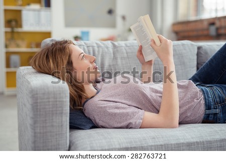 Profile of Young Brunette Woman Relaxing on Sofa and Reading Novel Book at Home