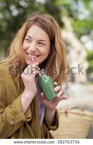 Close up Happy Blond Teen Girl Sipping a Bottle of Sweet Green Juice While Looking into Distance at her Back.