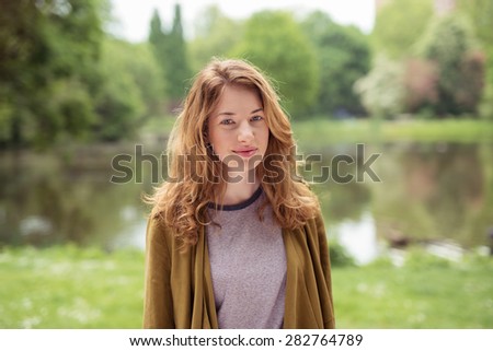 Half Body Shot of a Smiling Pretty Blond Teen Girl with Brown Cardigan, Standing at the Grassy Lakeside and Looking at the Camera.