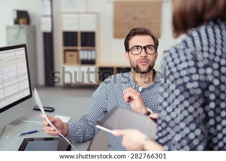 Young Handsome Office Man Talking to his Female Manager While Holding a Paper at his Working Table with Computer.