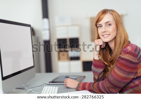 Friendly young redhead business woman sitting at her desk in the office in front of a desktop computer with a blank screen turning to smile at the camera