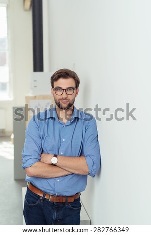Serious young man wearing casual blue shirt, brown leather belt, jeans, wristwatch and black frame eyeglasses while standing with folded arms leaning on a white wall and looking at camera