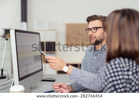 Proficient young male employee with eyeglasses and checkered shirt, explaining a business analysis displayed on the monitor of a desktop PC to his female colleague, in the interior of a modern office