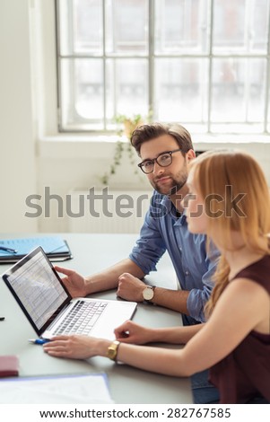Young Bearded Businessman Smiling at the Camera while Sitting at the Table Beside his Business Partner who is Busy at her Laptop.