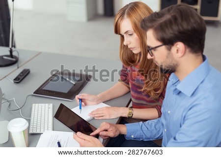 Young business partners working in the office sitting together at a desk using a tablet computer, man and woman with focus to the woman