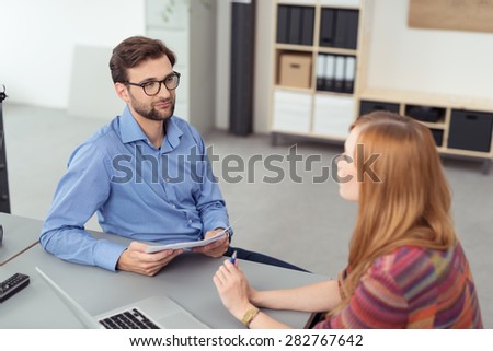 Businessman and woman having a discussion in the office as they relax at a desk facing one another, focus to the man