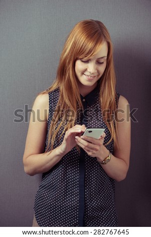 Attractive redhead young woman wearing a sleeveless shirt while smiling and using a smart phone for browsing internet, sending messages or networking, portrait with copy space on grey