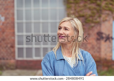 Smiling woman standing with folded arms looking up into the air watching something to the left of the frame with a brick building behind her