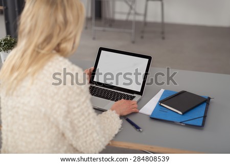 Blond woman working on a laptop computer as she sits at her desk, over the shoulder view of the blank white screen