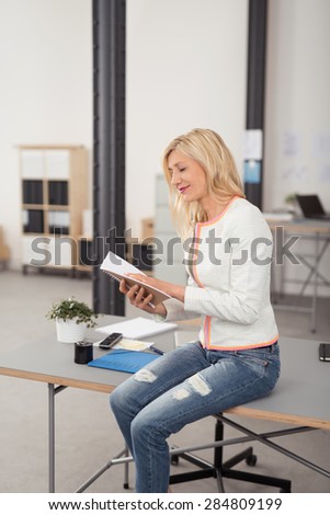 Smiling Blond Office Woman Sitting on a Gray Worktable While Reading Something in her Notebook