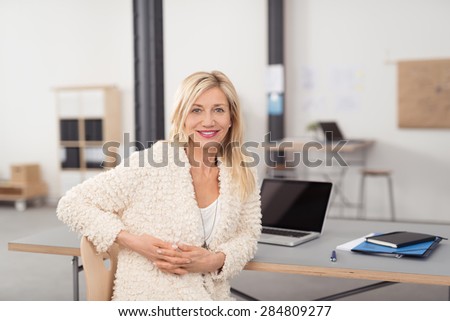 Optimistic Adult Office Lady in Off White Blazer, Sitting at her Table, with Laptop and Notes, with Hands Crossed and Looking at the Camera.