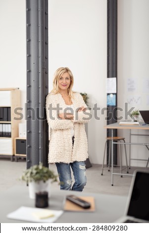 Confident Blond Office Woman Leaning Against Interior Metal Post with Arms Crossing Over her Stomach and Looking at the Camera.