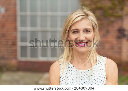 Beautiful blonde middle-aged woman, with medium-length hair and pink lipstick, wearing summer sleeveless dress with dotted pattern while smiling at camera, portrait in front of a building