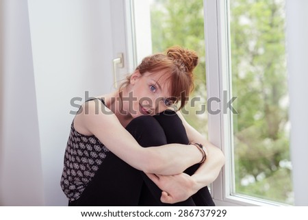 Smiling Cute Girl Hugging her Knees and Looking at the Camera While Sitting Near the Glass Window Inside the House.