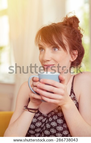 Close up Thoughtful Blond Young Woman Holding Cup of Coffee with her Two Hands and Looking Up with a Smiling Face.