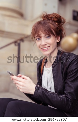 Young redhead woman with blue eyes, messy bun and jovial facial expression wearing a black leather jacket while using a mobile phone, sitting on the exterior stairs of a building