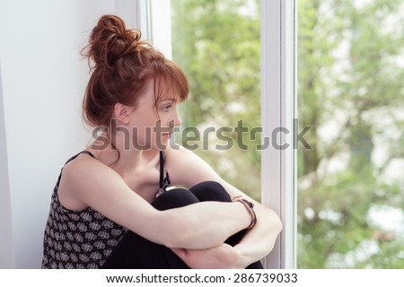 Sad young sitting hugging her knees on the window ledge woman staring out of the window at the garden outside