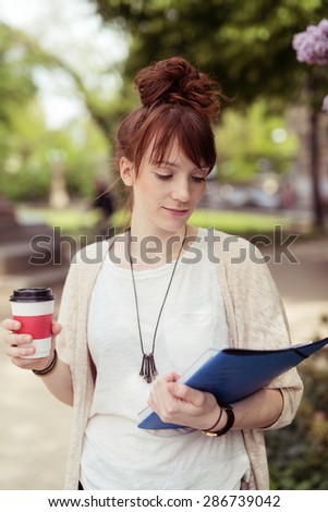 Pretty Young College Girl Holding a Cup of Coffee While Reading Something on the Book on her Other Hand Seriously.