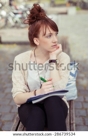 Pensive serious young woman sitting on a wooden park bench doing paperwork staring off to the right of the frame
