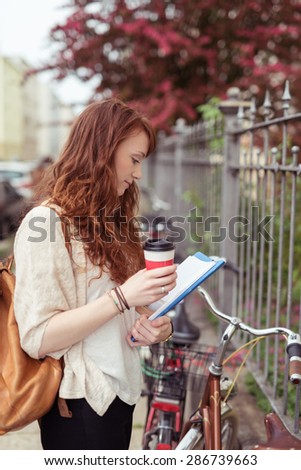 Attractive young student with long red hair standing on an urban street with a cup of takeaway coffee in her hand reading her notes alongside her bicycle