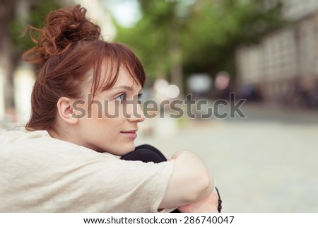Thoughtful young woman sitting hugging her knees as she sits staring thoughtfully into the air, profile view in an urban street