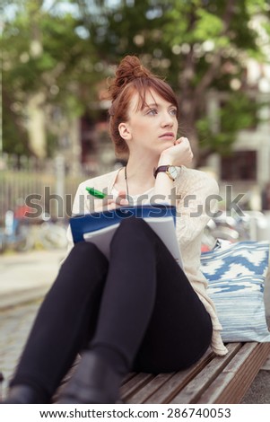 Pensive young redhead woman sitting on a wooden bench while getting inspired to write by the surrounding urban life, in a district with green vegetation