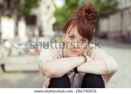 Young redhead woman sitting with her chin on her hands resting on her bent knees staring intently at the camera with a pensive expression