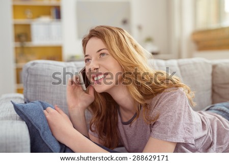 Young woman relaxing on her stomach on a sofa chatting on her mobile listening to the call with a delighted smile