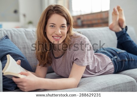 Pretty Teen Girl Holding a Novel Book, Lying on her Stomach on the Living Room Couch While Looking at the Camera with Feet Up