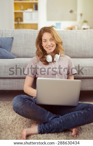 Happy young student working at home sitting cross-legged on the floor with her laptop computer and stereo headphones around her neck to listen to music smiling happily at the camera